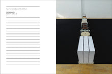 Load image into Gallery viewer, The inside of the book. On the left page is a title at the top, and then the remaining page is filled with horizontal lines stretching to the bottom. The right page features a photo of an artwork, containing many objects including tubes, rolls of fabric and paper, and stacks of notebooks. The objects are arranged standing up in a straight line that stretches away to the opposite wall.