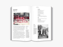 Load image into Gallery viewer, The inside of the book. The left page features the title “Bikyōtō 1968-74” and a black and white photo of a study with red text overlayed. The red text is a mixture of Japanese and English, with the largest piece of text being the word "revolution" in the centre. Below it is a paragraph of black text. The right page features black text and a black and white photo of a group of people walking through an office car park. Both pages are on a white background.