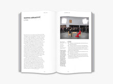 Load image into Gallery viewer, The inside of the book. The left page features the title "Marina Abramović" followed by several paragraphs of black text. The right page features a photo of a piece of performance art with a visitor and Marina Abramović sitting opposite eachother across a wooden table. Underneath the photo is a paragraph of black text. The background of both pages is white.