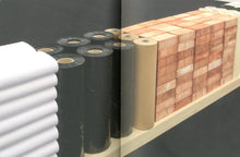 Load image into Gallery viewer, The inside of the book. Both pages feature a double-page spread of a photograph of an artwork. The piece contains many elements such as stacked bricks, rolls of paper, rolls of vinyl, and stacked papers, all arranged in a straight line.