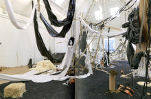 Load image into Gallery viewer, The inside of the book. Both pages feature a double-page spread of a photo of an artwork. The piece spans the entire gallery, and is made of piles of debris including bricks, straw, and wood, with some suspended from the ceiling. The room has black and white cloth looped from the rafters.